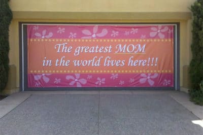 Large custom banner reading "The greatest MOM in the world lives here!!!" with floral design, showcasing event decoration potential for Mother's Day celebrations by 858 Graphics Inc.
