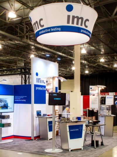 IMC branded trade show booth featuring a hanging sign, display screens, and promotional materials for productive testing, showcasing effective event visibility strategies.