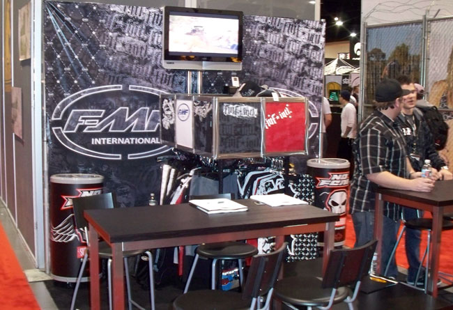 Trade show display booth featuring FMF International branding, showcasing merchandise, a monitor, and tables with chairs, set against a vibrant event backdrop.