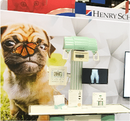 Pug dog with butterfly on nose in front of trade show display featuring veterinary equipment and branding for Henry Schein.