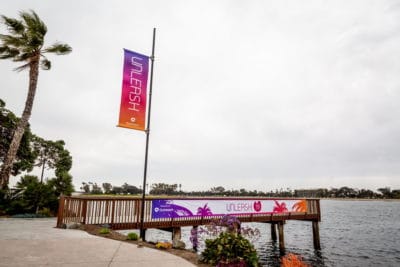 Colorful "UNLEASH" banner on a dock by a lake, showcasing custom branding for events, surrounded by palm trees and a scenic waterfront.