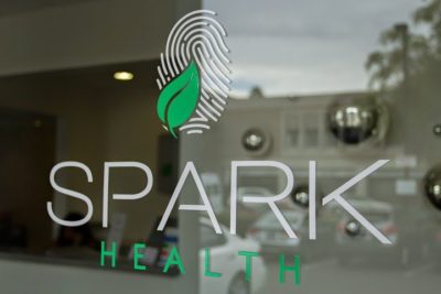 Window decal displaying "SPARK HEALTH" with a green leaf and fingerprint logo, promoting a welcoming atmosphere for local health services.