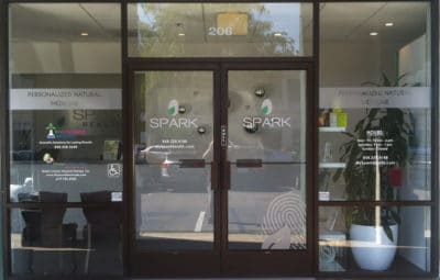 Glass entrance of a business with the name "SPARK" prominently displayed on the doors, featuring window decals that highlight personalized natural medical services, surrounded by a modern interior and greenery.