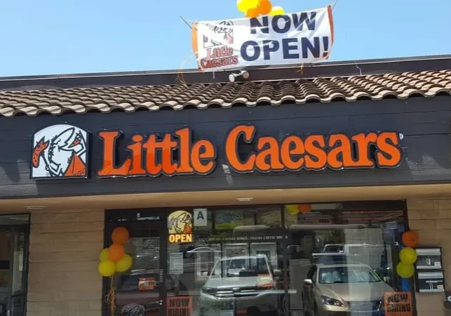 Little Caesars restaurant exterior with "NOW OPEN!" banner and colorful balloons.